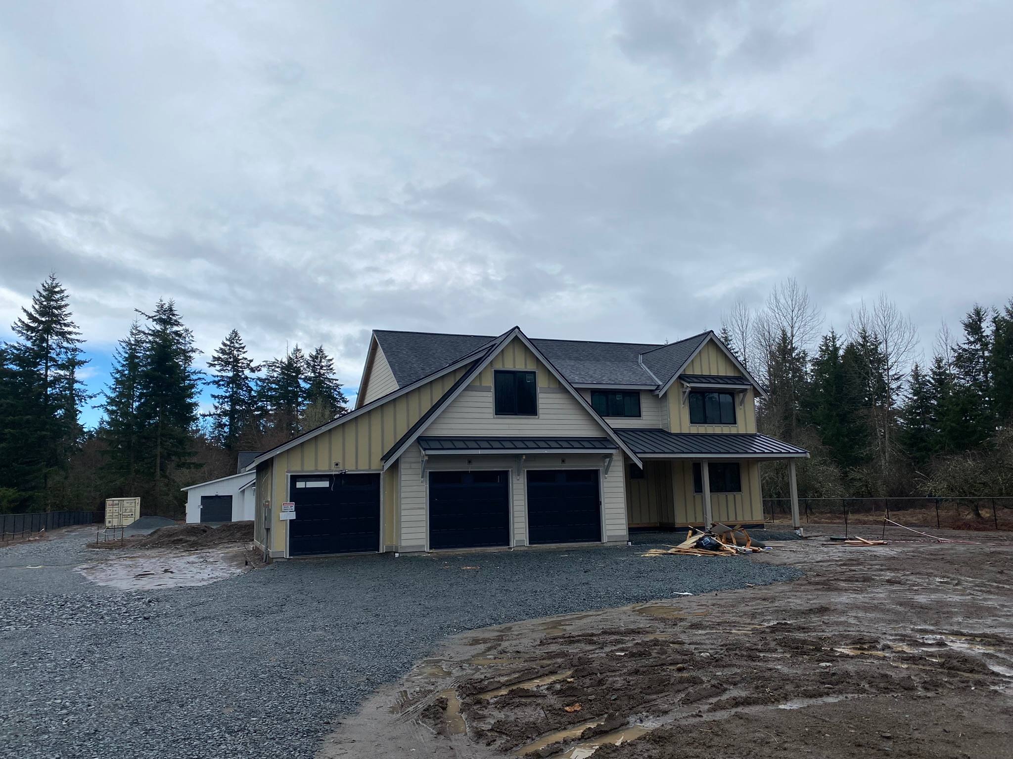 Two-story home with tan board and batten siding and black garage doors on a gravel lot